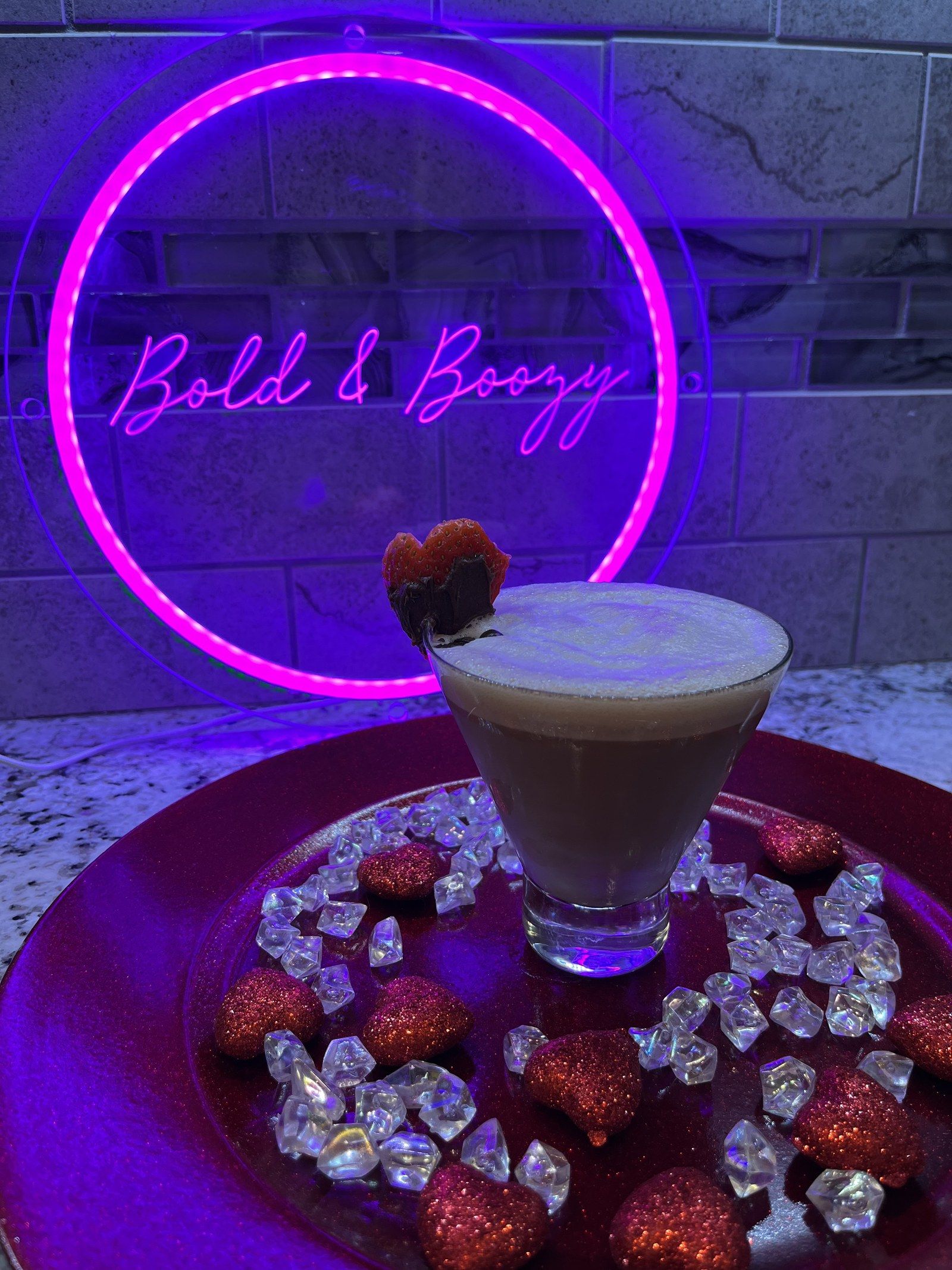 An espresso martini with a berry garnish in front of the glowing Bold & Boozy neon sign